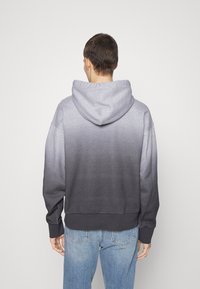 Šedá mikina s kapucí v přechodu odstínů, s měkkou texturou, kapsou na břiše a žebrovanými manžetami, přecházející od světlé po tmavou šedou.