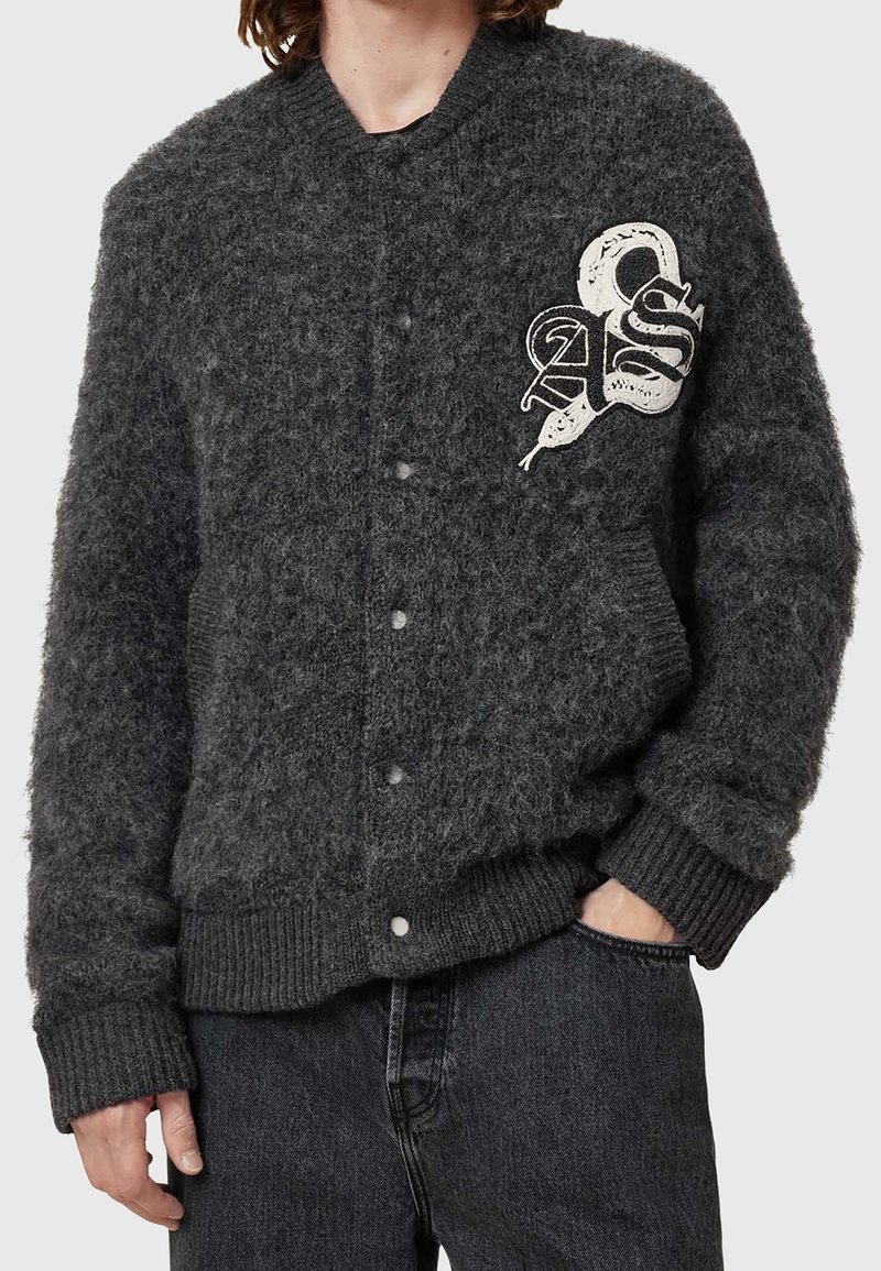 Personne portant un cardigan boutonné gris foncé duveteux avec un patch blanc représentant un serpent et des lettres sur la poitrine gauche, associé à un jean noir.