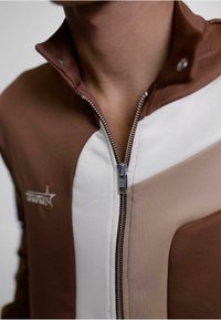 Brun og beige zip-up jakke med høy krage, med kontrasterende fargeblokker og metallglidelåsdetaljer. Logo på brystområdet.
