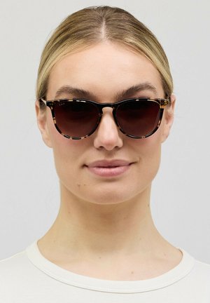 Vrouw met licht haar die donkere schildpadkleurige zonnebril draagt en een witte top, kijkt recht vooruit met een neutrale uitdrukking.