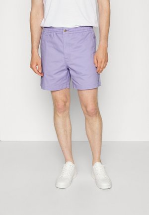 Person trägt hellviolette Shorts, weiße Sneakers und ein weißes Shirt und steht vor einem schlichten hellen Hintergrund.