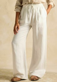 Biele nohavice s širokými nohavicami vyrobené z ľahkej tkaniny, s elastickým pásom a sťahovacou šnúrkou. Majú bočné vrecká a hladký povrch.