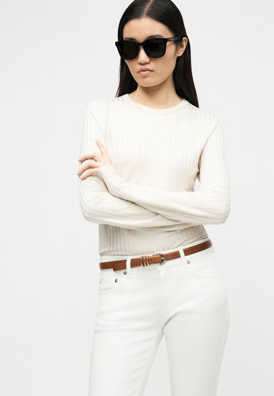 Γυναίκα με μακριά σκούρα μαλλιά που φοράει μαύρα γυαλιά ηλίου, κρεμ ριμπ μπλούζα με μακριά μανίκια, λευκό παντελόνι και καφέ ζώνη, όρθια με σταυρωμένα τα χέρια.