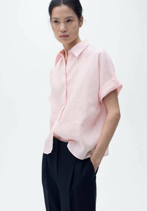Chemise en lin rose clair à manches courtes, devant boutonné et coupe décontractée, associée à un pantalon sombre et ajusté.