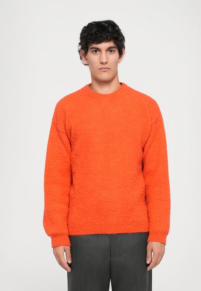 Joven de pie, de frente, vistiendo un suéter texturizado naranja brillante y pantalones oscuros grises, frente a un fondo liso.