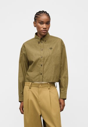 Femme aux cheveux tressés porte une chemise courte à motifs boutonnée et un pantalon beige taille haute devant un fond uni.