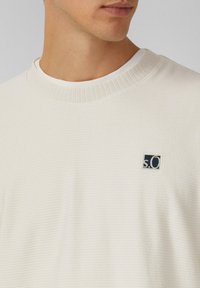 Ecru katoenen sweater met een geribde halslijn, gestructureerde stof en een klein marineblauw logo-opname op de linkerborst.