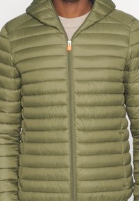 Giacca imbottita verde oliva con zip frontale, cappuccio e texture a righe orizzontali. Presenta un cursore arancione e una finitura in tessuto liscio.