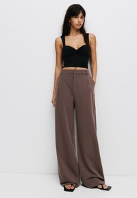 Černý přiléhavý crop top se širokými ramínky, v kombinaci s volnými hnědými kalhotami s vysokým pasem, které mají přední záhyby a boční kapsy.