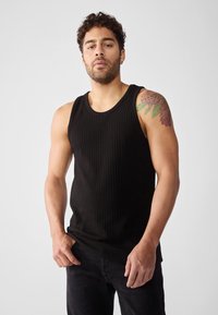Top in tessuto a costine nero con scollatura rotonda, caratterizzato da spalline larghe e un design aderente. Modello che indossa jeans neri e un tatuaggio visibile sul braccio.
