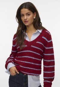 Frau mit langen dunklen Haaren, die Perlenohrringe trägt, einen rot-blauen gestreiften V-Ausschnitt-Pullover über einem hellblauen Button-Down-Hemd und dunklen Jeans.