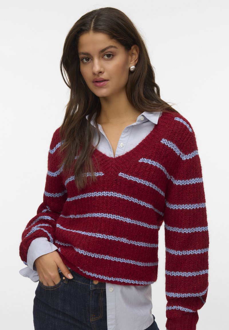 Frau mit langen dunklen Haaren, die Perlenohrringe trägt, einen rot-blauen gestreiften V-Ausschnitt-Pullover über einem hellblauen Button-Down-Hemd und dunklen Jeans.