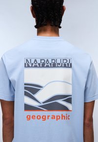 Light blue t-shirt featuring a graphic print of wavy lines in gray, with the text "NAPAPIJRI" above and "geographic" in orange below.