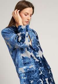 Blazer de terciopelo azul con flores, con un cuello estructurado, escote en V profundo y grandes bolsillos de parche. Presenta tela texturizada y patrones florales contrastantes.