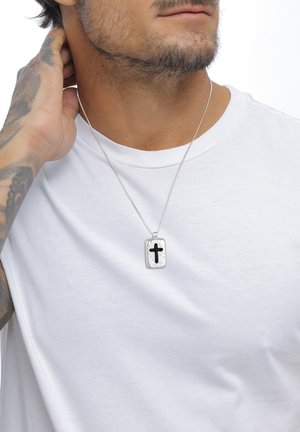 Sølvvedhæng med en struktureret overflade, der har et sort kors, hængende fra en tynd kæde, båret over en ensfarvet hvid t-shirt.