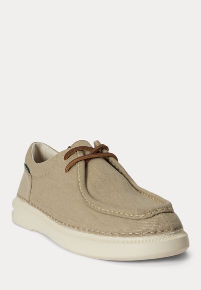 Bež platnena športna obutev z rjavimi vezalkami, belim podplatom in vidnim šivanjem, rahlo zasukana, da prikaže stran in sprednji del.