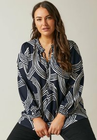 Marinebluse mit weißem Wellenmuster, ausgestattet mit einem Bindekragen und gerafften Manschetten. Strukturierter Stoff und lockerer Schnitt.