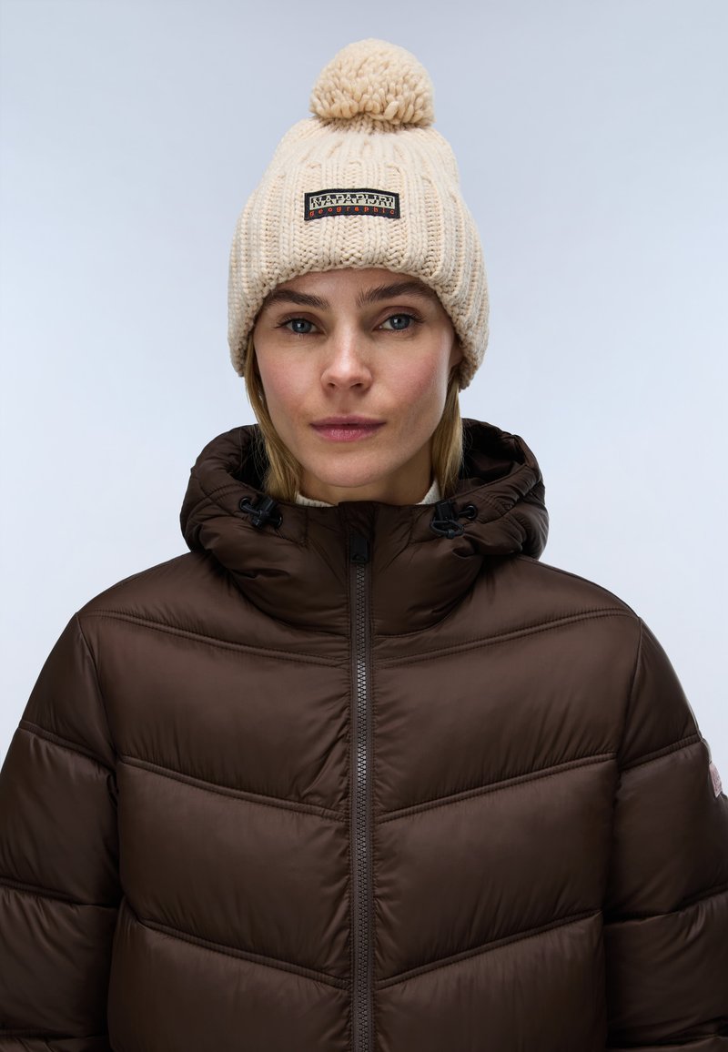 Femme portant un bonnet beige en tricot avec un pompon et une doudoune marron avec capuche, regardant vers l'avant sur un fond uni.