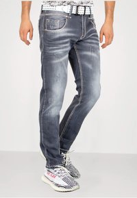 Graue, abgenutzte Jeans aus Denim mit kontrastierenden weißen Nähten, mehreren Taschen und leichter Ausbleichung. Kombiniert mit schwarz-weißen Sneakers mit Muster.