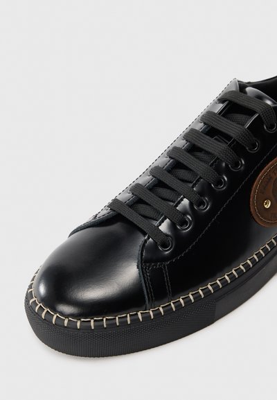 Schwarze Leder-Sneaker mit runder Spitze, flachen schwarzen Schnürsenkeln und braunem Etikett-Accent. Strukturierte Gummisohle mit sichtbarem Nähdetail.