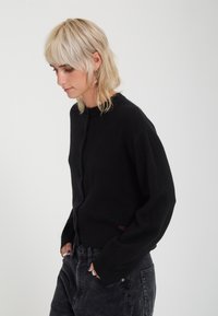 Jeune personne aux cheveux blonds courts portant un cardigan noir et un pantalon foncé, debout les mains dans les poches devant un fond uni.
