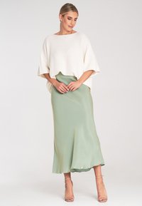 Weißes gestricktes Poncho-Oberteil kombiniert mit einem fließenden, hellgrünen Satinrock. Der Rock hat ein elegantes Finish und einen dezenten Glanz. Rosa Riemchen-Highheels runden das Outfit ab.