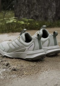 Ζευγάρι ανοιχτόχρωμα γκρι παπούτσια Adidas Terrex για τρέξιμο σε μονοπάτι, τοποθετημένα σε χαλίκι με πράσινη βλάστηση στο φόντο.