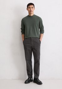 Dunkelgrüner Langarmpullover, kombiniert mit grauen gestreiften Hosen und schwarzen Schnürschuhen, vor einem schlichten Hintergrund stehend.