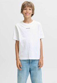 Usměvavý chlapec s světle hnědými vlasy, oblečený v bílém tričku s nápisem "CORE™ DEPT." a světle modrých džínách, stojící na jednobarevném pozadí.