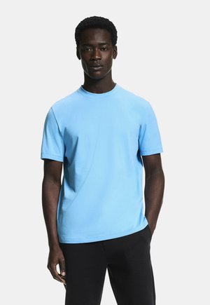 Rövid, göndör hajú férfi világoskék rövid ujjú ingben és fekete nadrágban, egyik kezét zsebében tartva áll.