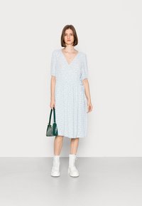 Vestido midi floral azul claro com mangas curtas em balão e decote em V. Modelo carrega uma bolsa verde escura e usa botas brancas com atacadores.