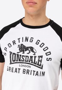 Balts T-krekls ar melniem raglan piedurknēm. Iekļauj tekstu "Sporting Goods Lonsdale London Great Britain" un lauvas grafiku nodilušā melnā printā.