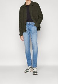 Tmavě zelená bunda z vlněné směsi s přední kapsou, černá košile, světle modré slim-fit džíny a vícebarevné tenisky se zelenými akcenty.