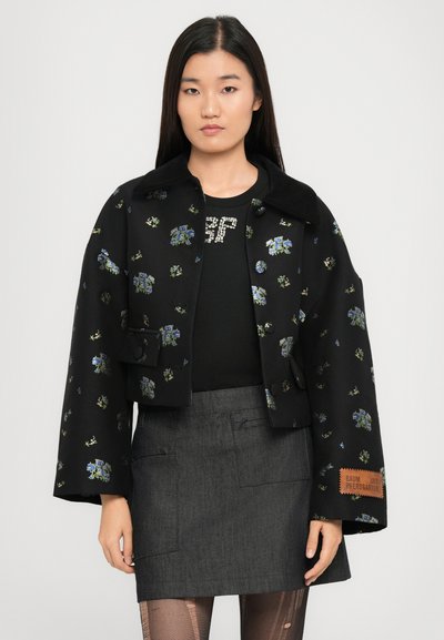 Κοντό μαύρο μπουφάν με λουλουδάτη κεντητική, φαρδιά μανίκια, κούμπωμα με κουμπιά και δερμάτινη ετικέτα με λογότυπο. Συνδυάζεται με γκρι τζιν φούστα και μαύρο πουκάμισο.