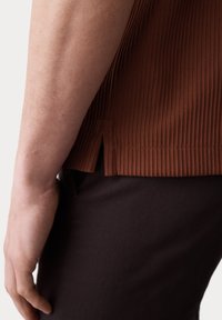 T-shirt marron côtelé avec ourlet et fentes sur les côtés, associé à un pantalon en tissu sombre. Peau visible sur la main et l'avant-bras.