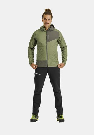 Mann mit Schnurrbart und Haarknoten, trägt grüne Kapuzenjacke, schwarze Hose und schwarze Schuhe mit leuchtend gelben Schnürsenkeln, steht vor weißem Hintergrund.