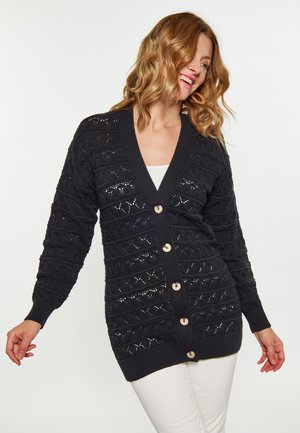 Frau lächelt und trägt eine schwarze, strukturierte Strickjacke mit großen Knöpfen über einem weißen Top und weißen Hosen vor einfarbigem Hintergrund.