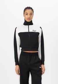 Veste zippée noire et blanche coupée avec col haut, présentant des manches longues et un patch logo ; associée à un pantalon de jogging noir assorti.