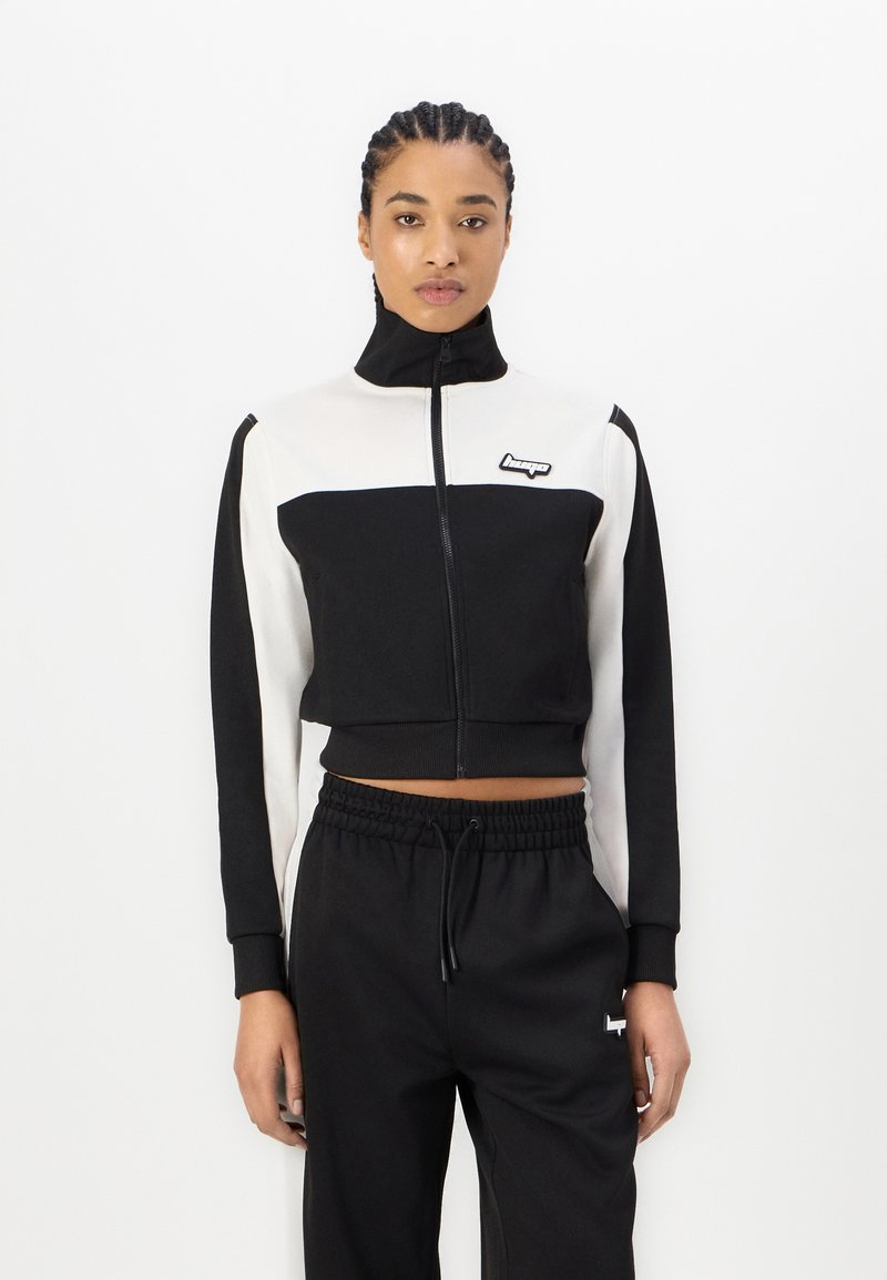 Giacca zip-up nera e bianca, con colletto alto, maniche lunghe e patch con logo; abbinata a pantaloni jogger neri coordinati.