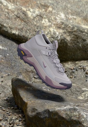 Светлосиви и лилави туристически обувки с връзки и подметка Vibram, висящи над скалист терен.