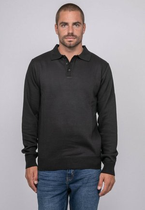 Homme aux cheveux courts et barbe, portant un polo à manches longues noir et un jean bleu, debout devant un fond uni.