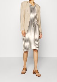 Blazer en lin beige sur une robe à rayures blanche cassée et marron, avec une taille ajustable. Des sandales à talons marron complètent la tenue.