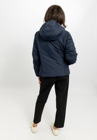 Giacca imbottita blu navy con cappuccio, caratterizzata da una superficie testurizzata e polsini elasticizzati, abbinata a pantaloni neri e scarpe di colore chiaro.