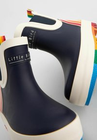 Bottes en caoutchouc navy à chevilles avec des accents crème et un talon à rayures multicolores de type arc-en-ciel. Présente l'étiquette "Little Bird by Jools Oliver".