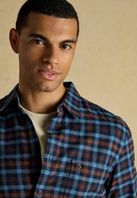 Zilā un brūnā kadrā veidota flaneļa krekls ar pogu apkakli un vienu krūšu kabatu. Mīksta tekstūra, apvienota ar bežīgu apakškrekli.