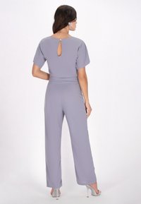 Lichtgrijze jumpsuit met korte mouwen en een detail met sleutelgat op de rug. Soepele textuur en een wijd broek ontwerp, gedragen met zilveren hoge hakken.