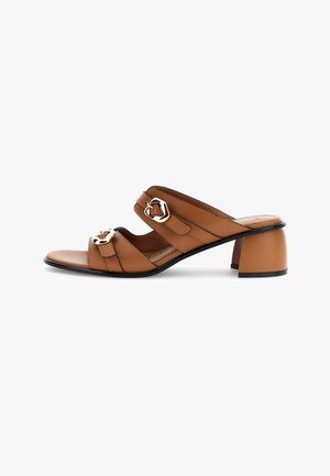 Ruskeat nahkaiset liukusäädettävät sandaalit, joissa on kaksi säädettävää remmiä kullanvärisillä soljilla ja matala paksu korko, jossa mustat pohjan reunat.