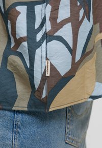 Gros plan sur un tissu à motif camouflage avec une étiquette « ORIGINALS » visible, porté sur un jean en denim bleu.