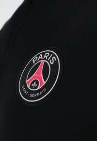 Schwarzer Sportbekleidungsstoff mit einem runden, gestickten Logo. Das Logo enthält den Text "PARIS," einen pinkfarbenen Eiffelturm und "SAINT-GERMAIN" in Weiß.