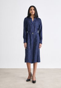 Abito camicia blu navy con maniche lunghe, motivo testurizzato, vita stretta con una cintura, lunghezza al ginocchio, abbinato a tacchi neri.